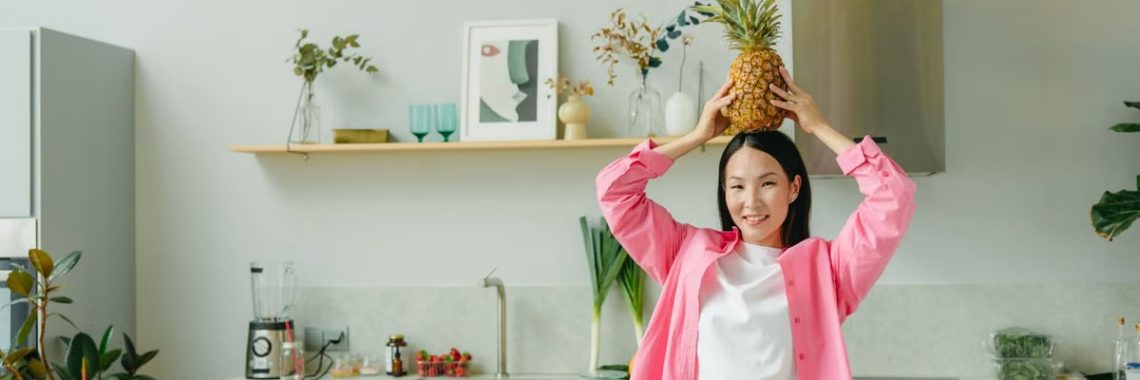 Asian woman in pink overshirt holding a pineapple in a modern kitchen with fresh produce and cheerful ambiance.
